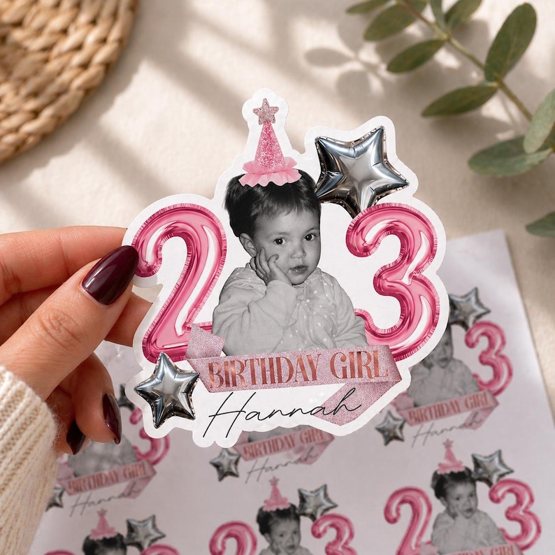 May include: A sticker featuring a black and white photo of a young child wearing a party hat, with the numbers "23" in pink balloon style. The words "BIRTHDAY GIRL Hannah" are printed on a pink ribbon.