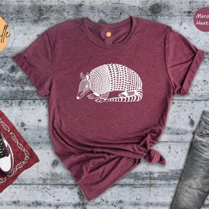May include: Maroon heather t-shirt with a white armadillo graphic. The shirt is styled with jeans, sneakers, and a red bandana. The text "Creativille" is in a circle in the upper left corner, and "Maroon Heather" is in a circle in the upper right.