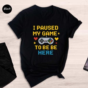 May include: Black t-shirt with the text "I paused my game to be here" in a pixelated font. The text is surrounded by colourful pixelated hearts and a pixelated game controller.