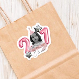 May include: A sticker on a brown paper bag features a black and white photo of a young girl wearing a party hat. Pink balloon numbers '21' and silver star balloons surround the photo. The text 'BIRTHDAY GIRL Olivia' is on a pink banner.