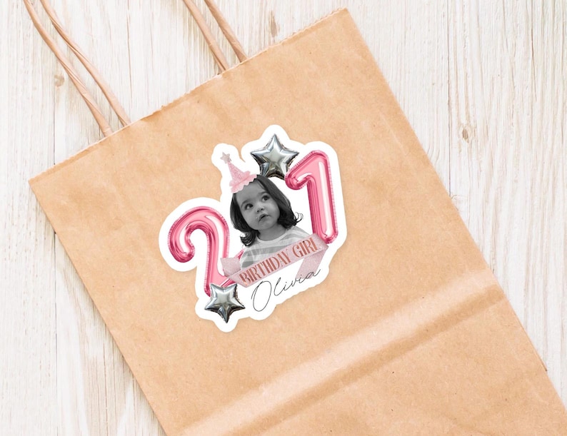 May include: A sticker on a brown paper bag features a black and white photo of a young girl wearing a party hat. Pink balloon numbers '21' and silver star balloons surround the photo. The text 'BIRTHDAY GIRL Olivia' is on a pink banner.