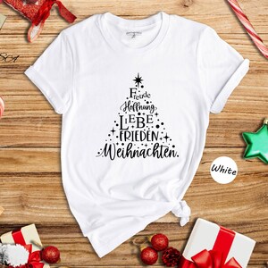 May include: White t-shirt with a Christmas tree design made of text in black. The text includes words like "Freude", "Hoffnung", "Liebe", "Frieden", and "Weihnachten". The shirt is on a wooden surface with Christmas decorations.