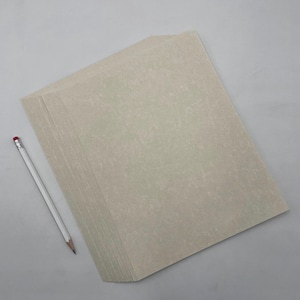 May include: A stack of beige-colored paper with a textured surface. A white pencil with a red eraser rests beside the paper. The paper and pencil are on a gray surface.