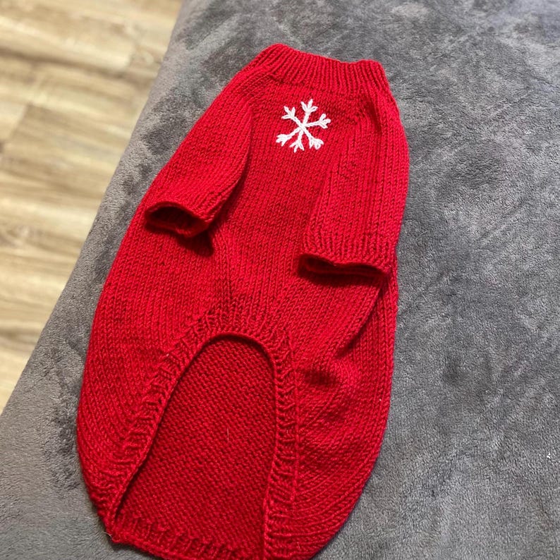 Pu&ograve; includere: Un maglione lavorato a maglia di colore rosso acceso per animali domestici, dotato di colletto a coste e un motivo a fiocco di neve bianco ricamato al centro della parte superiore della schiena. Presenta maniche corte e un orlo curvo.