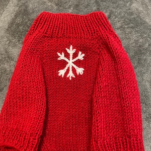 Pu&ograve; includere: Un maglione lavorato a maglia rosso brillante con colletto a coste e un disegno a fiocco di neve bianco ricamato al centro del petto. Il capo &egrave; realizzato con un motivo a maglia verticale su tutto il corpo e le maniche, con una finitura.