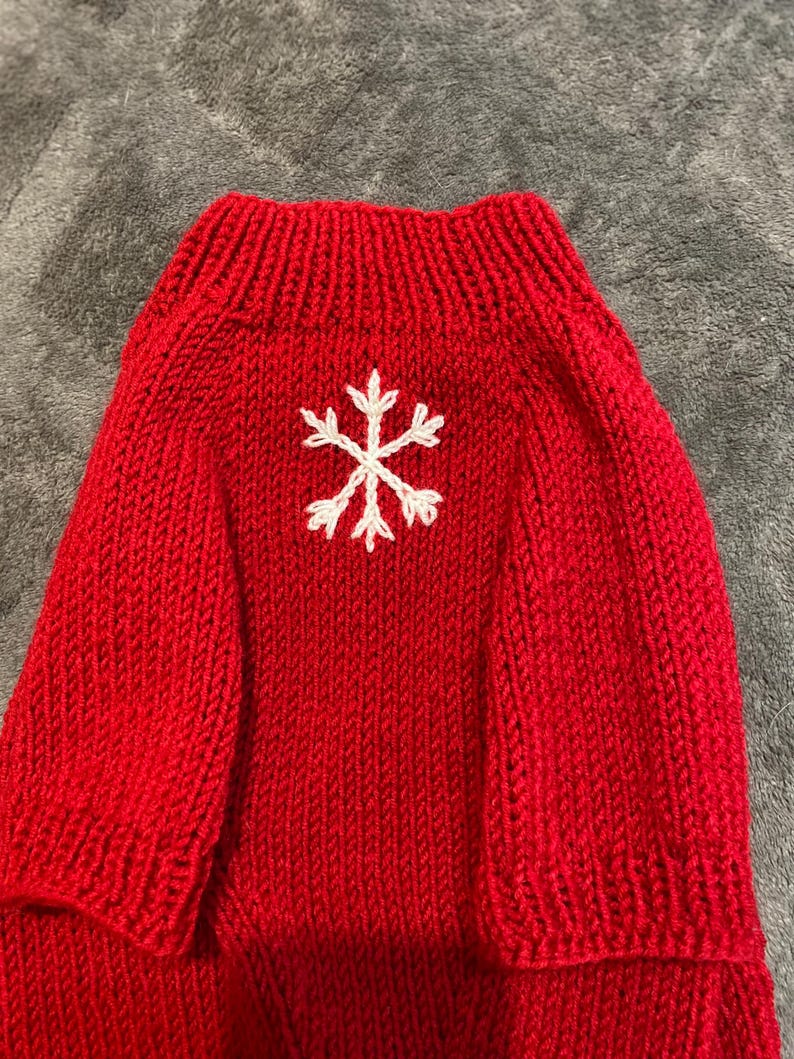 Pu&ograve; includere: Un maglione lavorato a maglia rosso brillante con colletto a coste e un disegno a fiocco di neve bianco ricamato al centro del petto. Il capo &egrave; realizzato con un motivo a maglia verticale su tutto il corpo e le maniche, con una finitura.