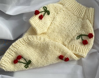 May include: A cream-colored knitted sweater and matching hat set. The sweater has short sleeves and a ribbed neckline. Both pieces are adorned with embroidered red cherries and green stems.