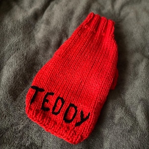 Pu&ograve; includere: Un maglione lavorato a maglia di colore rosso brillante con colletto a coste e il nome TEDDY ricamato in grassetto con lettere maiuscole nere nella parte inferiore. Il capo presenta un motivo a maglia verticale e un design senza maniche.