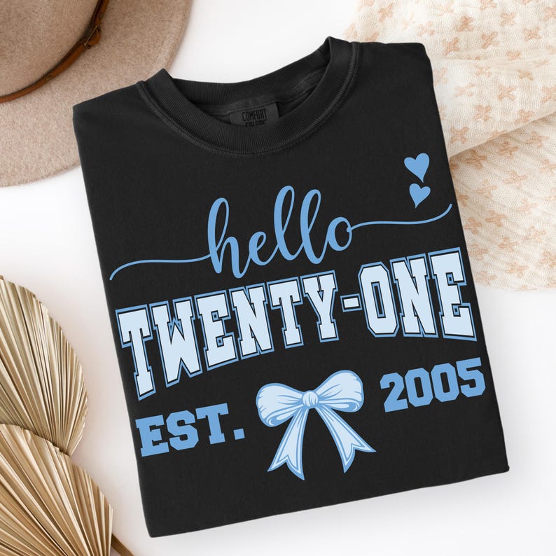 May include: Black crew neck t-shirt with the text "hello TWENTY-ONE EST. 2005" in light blue. The design includes two small blue hearts and a bow. The shirt is folded and displayed on a white surface.