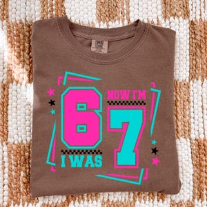May include: A brown t-shirt with the text "I WAS 67 NOW I'M" in a retro design. The numbers 6 and 7 are in bright pink and teal, with a black and white checkered pattern. The shirt has a crew neck and short sleeves.