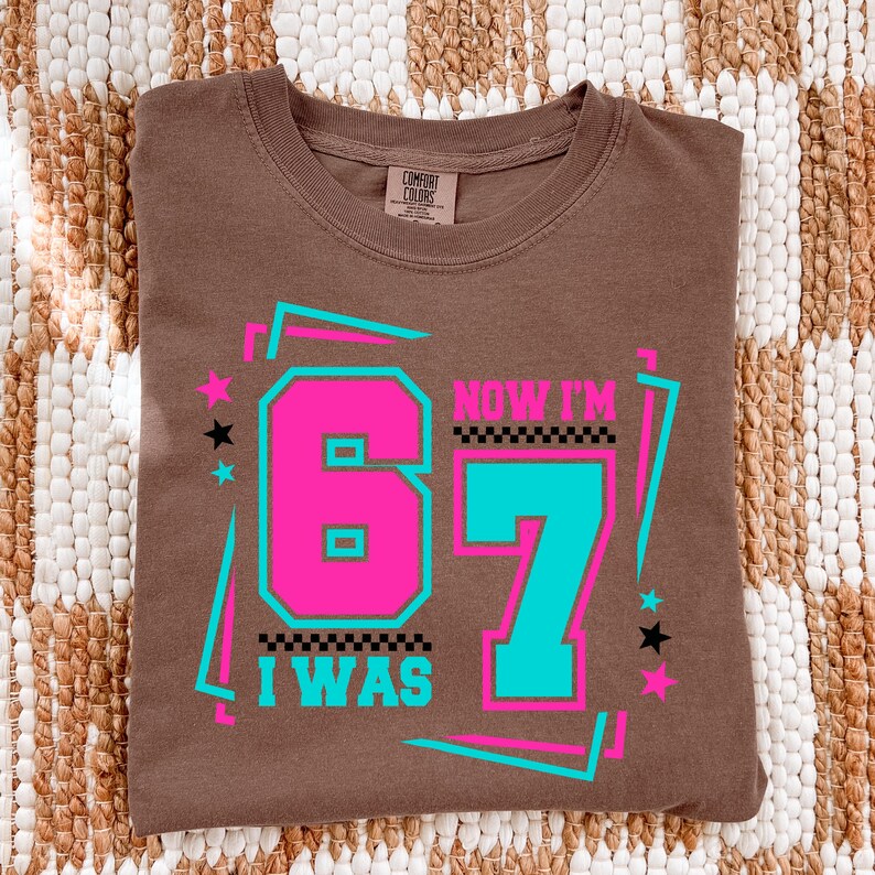 May include: A brown t-shirt with the text "I WAS 67 NOW I'M" in a retro design. The numbers 6 and 7 are in bright pink and teal, with a black and white checkered pattern. The shirt has a crew neck and short sleeves.