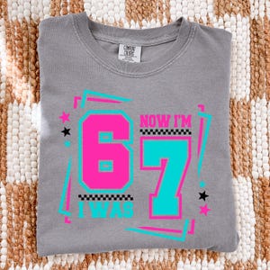 May include: Gray Comfort Colors t-shirt with the text "NOW I'M 67 I WAS" in a retro design. The number 67 is in pink and teal, with stars and a checkered pattern. The shirt is folded on a woven surface.