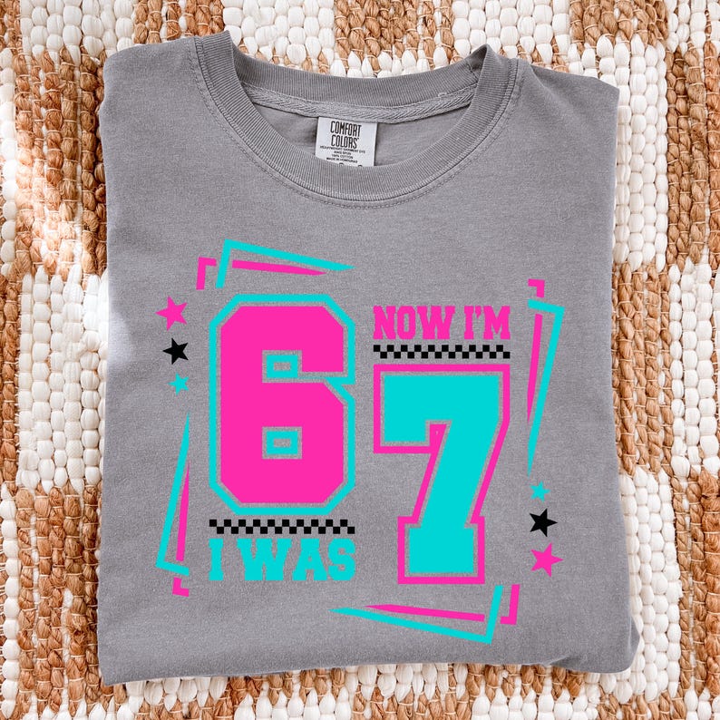 May include: Gray Comfort Colors t-shirt with the text "NOW I'M 67 I WAS" in a retro design. The number 67 is in pink and teal, with stars and a checkered pattern. The shirt is folded on a woven surface.