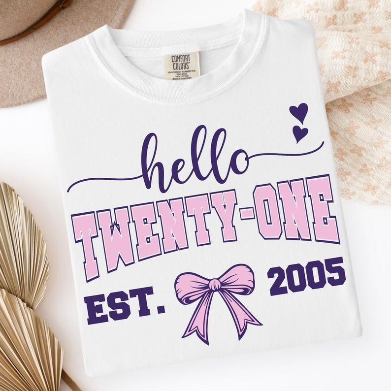 May include: White t-shirt with the text "hello TWENTY-ONE" in purple and pink, along with a pink bow and the year "2005". The shirt has a crew neck and a tag on the upper left side.