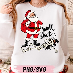 May include: A white long-sleeve sweatshirt featuring a graphic of Santa Claus standing over a fallen skeleton wearing a witch hat. The text "Well, shit..." is printed in black script next to the figures. The design includes red, black, and white colors.
