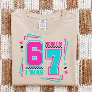 May include: A beige sweatshirt with the text "NOW I'M 67 I WAS" in a retro design. The number 6 is pink, and the number 7 is teal, with black and pink stars and lines. The sweatshirt is folded.