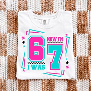 May include: White t-shirt with a graphic design that says "I WAS 67 NOW I'M 67" in pink and teal. The design includes stars and a checkered pattern. The shirt is folded on a woven surface.
