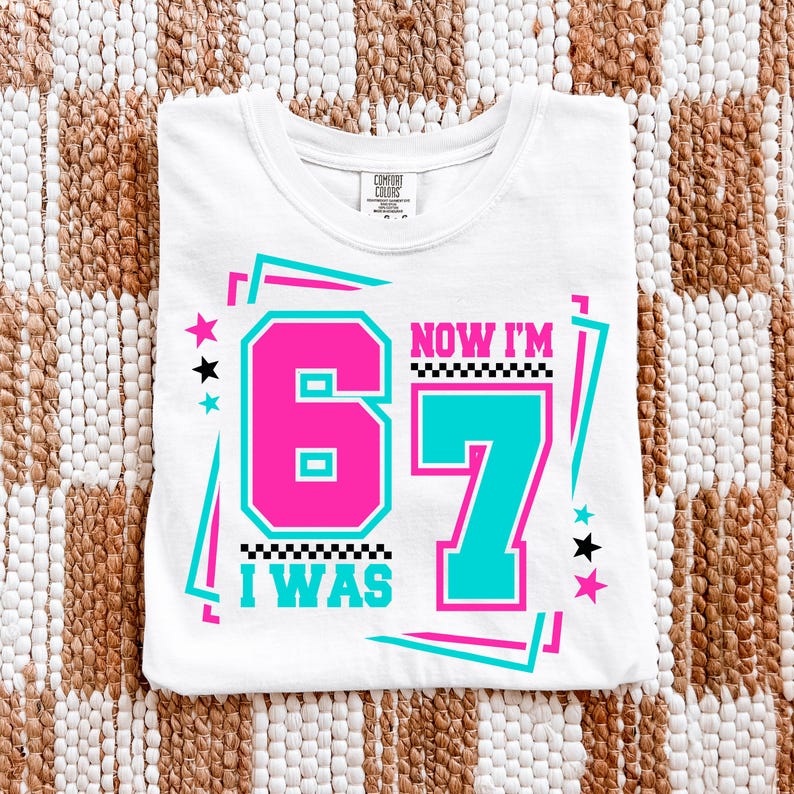 May include: White t-shirt with a graphic design that says "I WAS 67 NOW I'M 67" in pink and teal. The design includes stars and a checkered pattern. The shirt is folded on a woven surface.