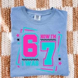 May include: A light blue t-shirt with the text "NOW I'M 67 I WAS" in a retro design. The number 6 is hot pink, and the number 7 is teal. The shirt has a crew neck and is made of a soft material.