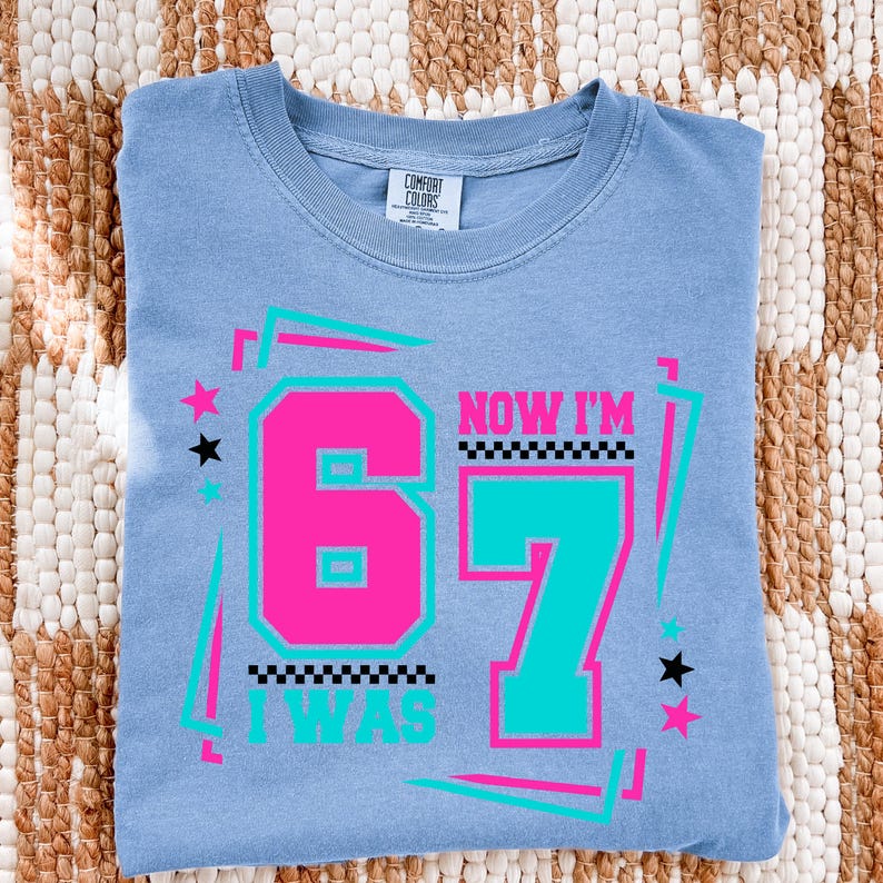 May include: A light blue t-shirt with the text "NOW I'M 67 I WAS" in a retro design. The number 6 is hot pink, and the number 7 is teal. The shirt has a crew neck and is made of a soft material.