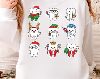 May include: A white long-sleeve sweatshirt featuring a graphic print of nine cartoon teeth characters dressed in various holiday-themed outfits, including Santa hats, reindeer antlers, and festive accessories, with the text PNG/SVG at the bottom.