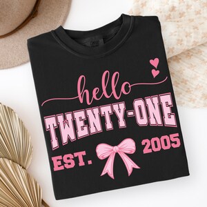 May include: Black t-shirt with the text "hello TWENTY-ONE EST. 2005" in pink. The design includes a pink bow and two pink hearts. The shirt is folded and displayed on a white surface.