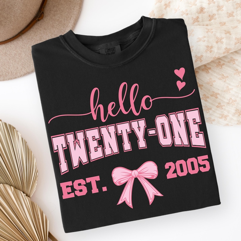 May include: Black t-shirt with the text "hello TWENTY-ONE EST. 2005" in pink. The design includes a pink bow and two pink hearts. The shirt is folded and displayed on a white surface.