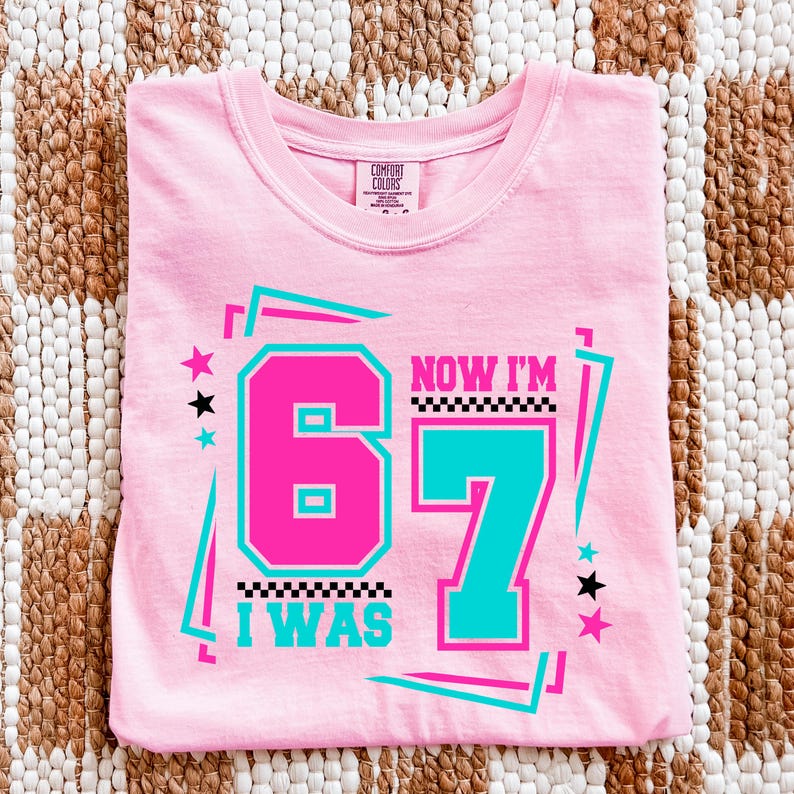May include: A pink t-shirt with the text "I WAS 67 NOW I'M 67" in a retro design. The numbers are in pink and teal, with black and white accents and star graphics. The shirt is made of a soft material.