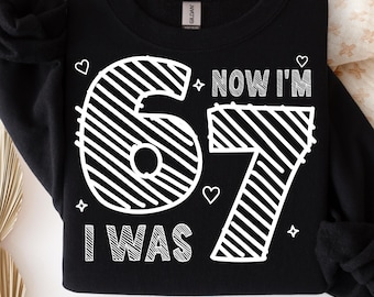 May include: Black sweatshirt with the text "I WAS 67 NOW I'M 67" in white, with heart and star accents. The bottom of the sweatshirt has the text "PNG/SVG" in pink.