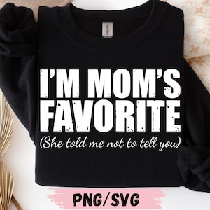 May include: Black sweatshirt with the text "I'M MOM'S FAVORITE (She told me not to tell you)" in white block letters. The bottom of the image has the text "PNG/SVG" in pink.