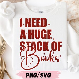 May include: White sweatshirt with the text "I NEED A HUGE STACK OF Books" in a bold, red font. The design includes decorative star accents. The bottom of the image includes the text "PNG/SVG".