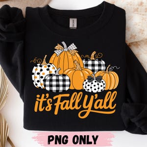 May include: A black crewneck sweatshirt featuring a graphic of six pumpkins in orange, black, and white with polka dot and buffalo plaid patterns. The text "it's Fall Y'all" is printed in orange below the pumpkins. A Gildan brand tag is visible.