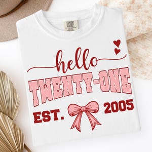 May include: White crew neck t-shirt with the text "hello TWENTY-ONE EST. 2005" in red and pink. The design includes two small red hearts and a pink bow. The shirt is folded.