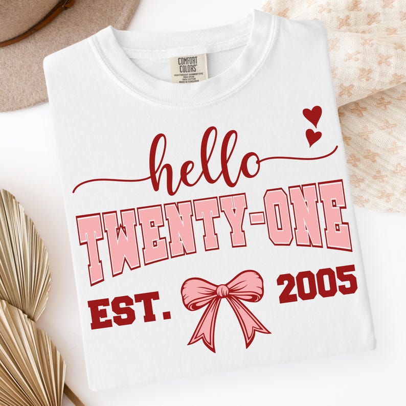 May include: White crew neck t-shirt with the text "hello TWENTY-ONE EST. 2005" in red and pink. The design includes two small red hearts and a pink bow. The shirt is folded.