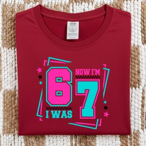 May include: A maroon t-shirt with the text "NOW I'M 67 I WAS" in a retro design. The number 6 is pink, the number 7 is teal, and the text is outlined in black. The shirt is folded on a patterned surface.