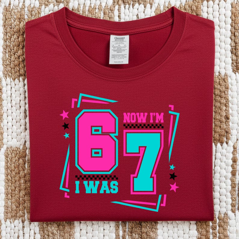 May include: A maroon t-shirt with the text "NOW I'M 67 I WAS" in a retro design. The number 6 is pink, the number 7 is teal, and the text is outlined in black. The shirt is folded on a patterned surface.