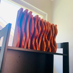 May include: A set of wooden wall decor pieces with a wavy, organic design. The pieces are stacked on a shelf and are made of a reddish-brown wood.