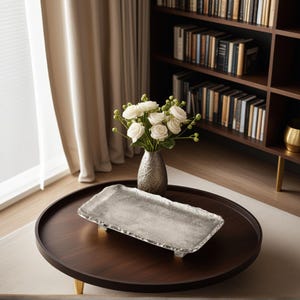 May include: A silver rectangular tray with a textured surface and decorative edges sits on a dark brown round coffee table. A vase with white roses and a bookshelf are in the background. The table has gold-coloured legs.