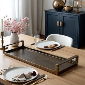 May include: A rectangular, gold-toned serving tray with two handles. The tray is on a wooden table with a white plate and cutlery. There are pink flowers in a white vase on the table.