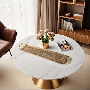 May include: A circular white marble coffee table with a gold base. A long, rectangular gold tray with handles is on the table, alongside a small gold vase of flowers and a magazine. The table is in a living room.