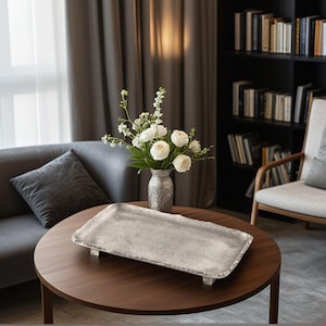 May include: A silver rectangular tray with a textured surface and short feet rests on a dark brown wooden table. A vase with white flowers sits behind the tray. The room has a grey sofa, a bookshelf, and a window with grey curtains.