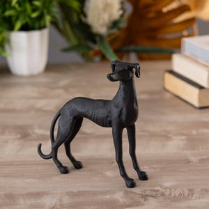 May include: Black metal sculpture of a greyhound dog standing on a wooden surface.