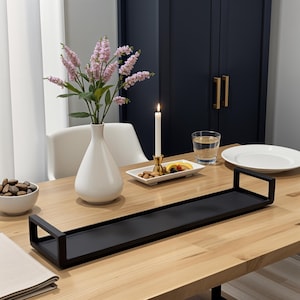 May include: A black metal serving tray with a raised edge sits on a light wood table. A white vase with pink flowers, a white plate with nuts and a lit candle are on the table.