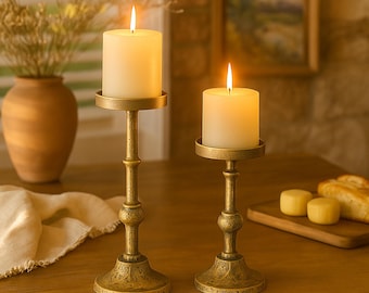 May include: Two lit ivory pillar candles sit atop antique gold candle holders. The candle holders have a detailed, ornate design and are placed on a wooden surface. A vase with dried flowers is in the background.