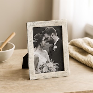 May include: A rectangular photo frame with a distressed, off-white finish. The frame holds a black and white photograph of a couple. The frame is propped up on a wooden surface, with a bowl and a folded blanket in the background.