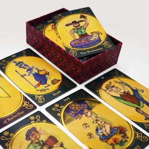Op de afbeelding: Een set tarotkaarten met kleurrijke illustraties en gouden accenten. De kaarten tonen een verscheidenheid aan personages en symbolen, waaronder een skelet, een vlinder en een man met een hoge hoed. De kaarten zijn in een waaier geordend, met de doos op de achtergrond.