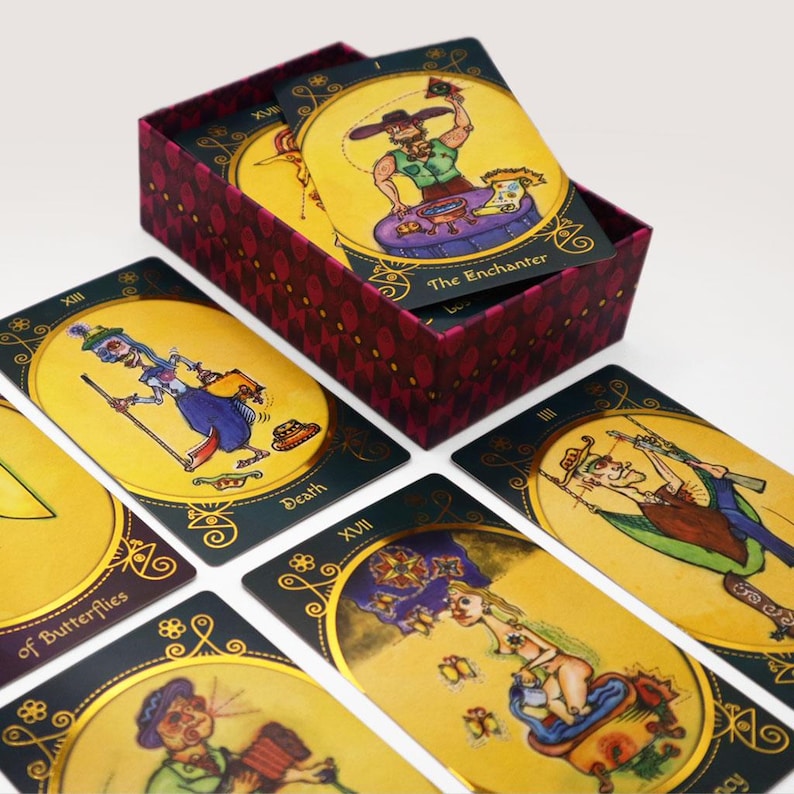 Op de afbeelding: Een set tarotkaarten met kleurrijke illustraties en gouden accenten. De kaarten tonen een verscheidenheid aan personages en symbolen, waaronder een skelet, een vlinder en een man met een hoge hoed. De kaarten zijn in een waaier geordend, met de doos op de achtergrond.