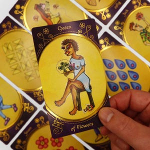 Op de afbeelding: Een tarotkaart met een paarse achtergrond en gouden rand. De kaart toont een vrouw met een bruine huid die op een kruk zit en een boeket bloemen vasthoudt. De kaart is gelabeld met "Koningin van Bloemen".