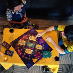 Op de afbeelding: Een tarotkaartlezing met een paarse doek met gouden accenten en vijf kaarten die op een gele tafelkleed zijn gelegd. Twee mensen zitten aan de tafel, een wijst naar een kaart en de ander kijkt naar de kaarten.