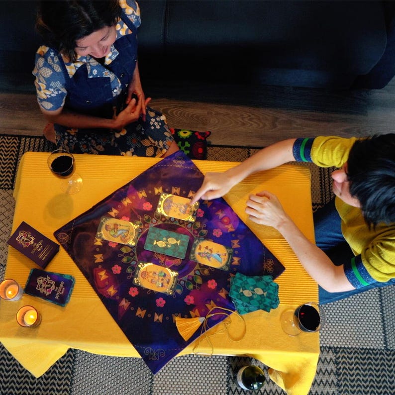 Op de afbeelding: Een tarotkaartlezing met een paarse doek met gouden accenten en vijf kaarten die op een gele tafelkleed zijn gelegd. Twee mensen zitten aan de tafel, een wijst naar een kaart en de ander kijkt naar de kaarten.